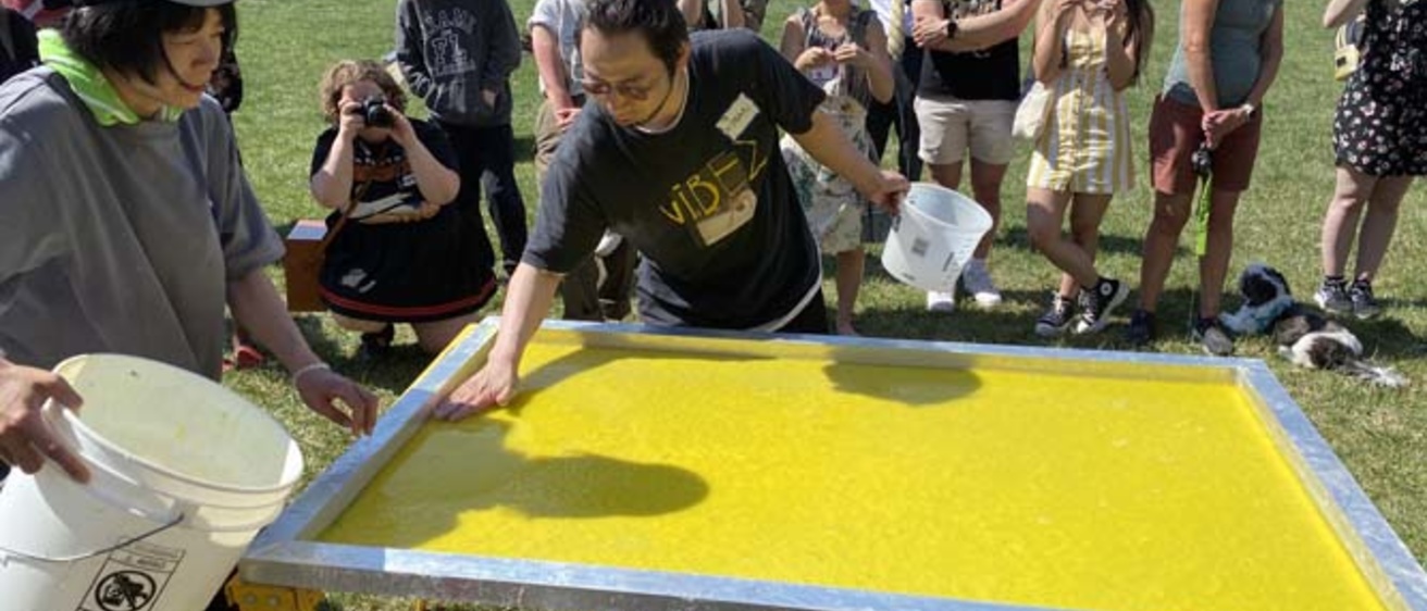 A group of people stand around a papermaking screen with yellow pulp drying