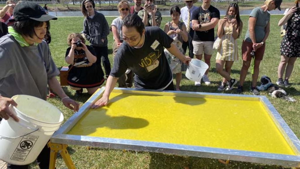 A group of people stand around a papermaking screen with yellow pulp drying